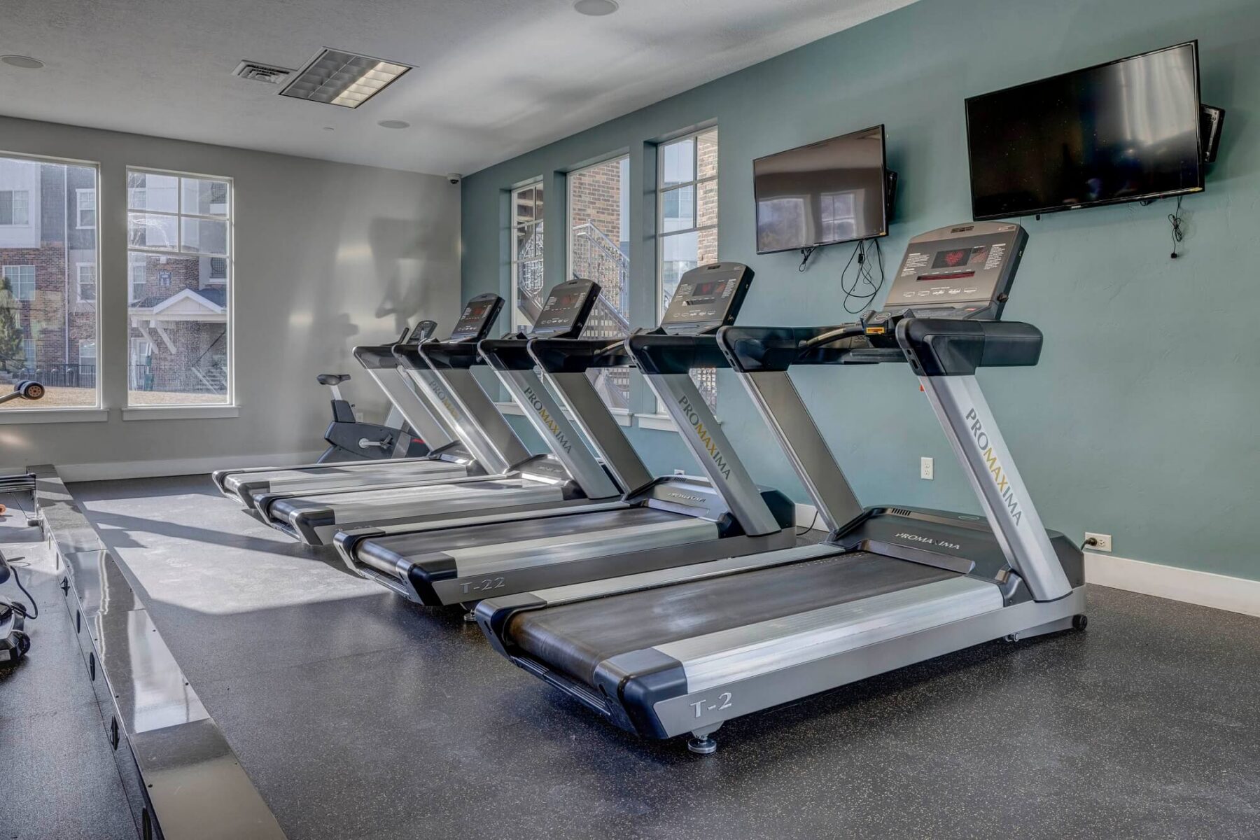 A modern gym with four treadmills lined up in front of teal walls, two wall-mounted TVs above them, large windows letting in natural light, and an exercise bike in the background.